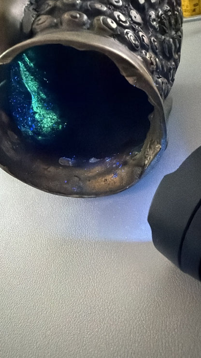 UV-Aufnahme eines antiken thailändischen bronzenen Buddha‑Kopfs mit interner Fluoreszenz und alter Patina.